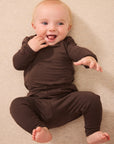 A smiling baby lying on a beige textured blanket, wearing a matching dark brown long-sleeve top and pants set.