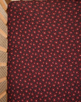 A close-up of a crib mattress fitted sheet with a dark red and black floral pattern, shown inside a wooden crib.