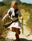 A young girl wearing a cream ribbed dress and brown boots runs along a dirt path outdoors, holding a large yellow flower cutout and a gray stuffed animal.