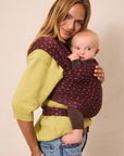 A woman wearing a lime green sweater and blue jeans carries a baby in a dark red baby wrap with a small red floral pattern, set against a plain beige background.