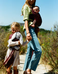 A woman carrying a baby in a dark red patterned baby carrier wrap walks outdoors on a dirt path with a young girl beside her holding a gray stuffed bunny.