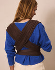 A woman shown from the back wearing a dark brown wrap-style baby carrier or shoulder harness over a royal blue sweater and cream pants, against a plain beige studio background.