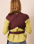 A woman viewed from behind wearing a wrap-style baby carrier or shoulder wrap in dark red with a small repeating floral pattern, worn over a yellow-green top and blue jeans against a plain beige studio background.