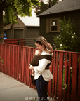A woman wearing a white shirt and dark jeans carries a baby in a brown wrap carrier outdoors on a sidewalk next to a red wooden fence.