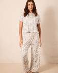 A woman modeling a matching two-piece pajama set with a short-sleeve top and wide-leg pants, featuring a light beige base with a blue toile pattern, set against a plain beige studio backdrop.