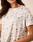 A close-up of a woman wearing a short-sleeve crewneck t-shirt with a blue toile de Jouy pattern on a cream-colored fabric, set against a warm beige background.