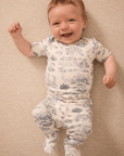 Smiling baby wearing cream romper with blue pastoral scene print, lying on beige textured fabric background