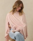 Woman wearing a light pink nursing cover while breastfeeding a baby in light pink socks, sitting on a wooden stool.