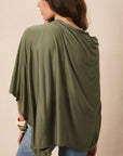 A woman wearing an olive green draped poncho-style top with blue jeans, shown from the back against a plain beige studio background.