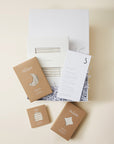 Baby gift set with beige striped sleeper, swaddle, and hat in brown packaging, displayed in white box with shredded paper.