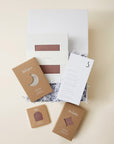 Brown baby swaddle, sleeper, and hat packaged in cardboard boxes with white shredded filler in an open white gift box.