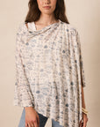 A woman wearing a draped, lightweight poncho-style top with a blue floral toile pattern on a cream background, paired with light-wash blue jeans, set against a plain beige studio background.