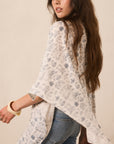 A woman wearing a loose, off-white floral kimono-style top with blue jeans, photographed against a plain beige studio background.