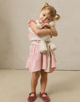 Toddler girl in pink dress and maroon shoes hugging cream plush toy secured with floral wrap carrier.