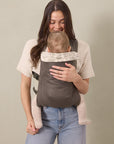 Woman wearing a dark gray baby carrier with beige patterned head support holding a baby facing inward.