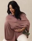Woman wearing dusty rose nursing cover while holding a baby dressed in olive green pants and white top