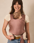 A woman wearing a dusty rose baby carrier wrap with a baby inside, standing against a plain beige background.