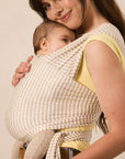 A woman wearing a beige and white gingham baby wrap carrier with a baby inside, styled over a yellow top against a plain beige background.