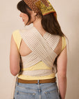 A woman shown from the back wearing a yellow sleeveless top with a beige and white striped wrap-style back panel, blue jeans, and a patterned brown headscarf, against a plain beige studio background.