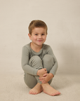 Young boy wearing a mist pointelle sleep set sitting on a beige surface with a plain background