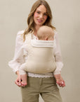 Woman wearing beige baby carrier holding baby facing inward, dressed in white blouse and olive green pants.