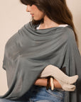 A woman wearing a draped sage green shawl or wrap over her shoulders and around her arms, with blue jeans and a white top visible. She is posed against a plain beige studio background.