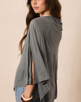 A woman wearing a draped, oversized gray top with wide sleeves and side slits, paired with blue jeans, photographed against a plain beige studio background.