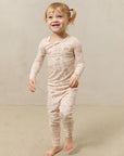 Toddler girl wearing beige long-sleeve top and pants set with light brown scenic pattern, standing barefoot on beige carpet.