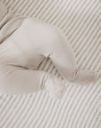 A close-up of a baby's legs and feet wearing a soft, light beige footed sleeper or bootie, lying on a striped beige and white mattress.