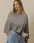 Black and beige houndstooth poncho on model sitting wearing light blue jeans against beige background