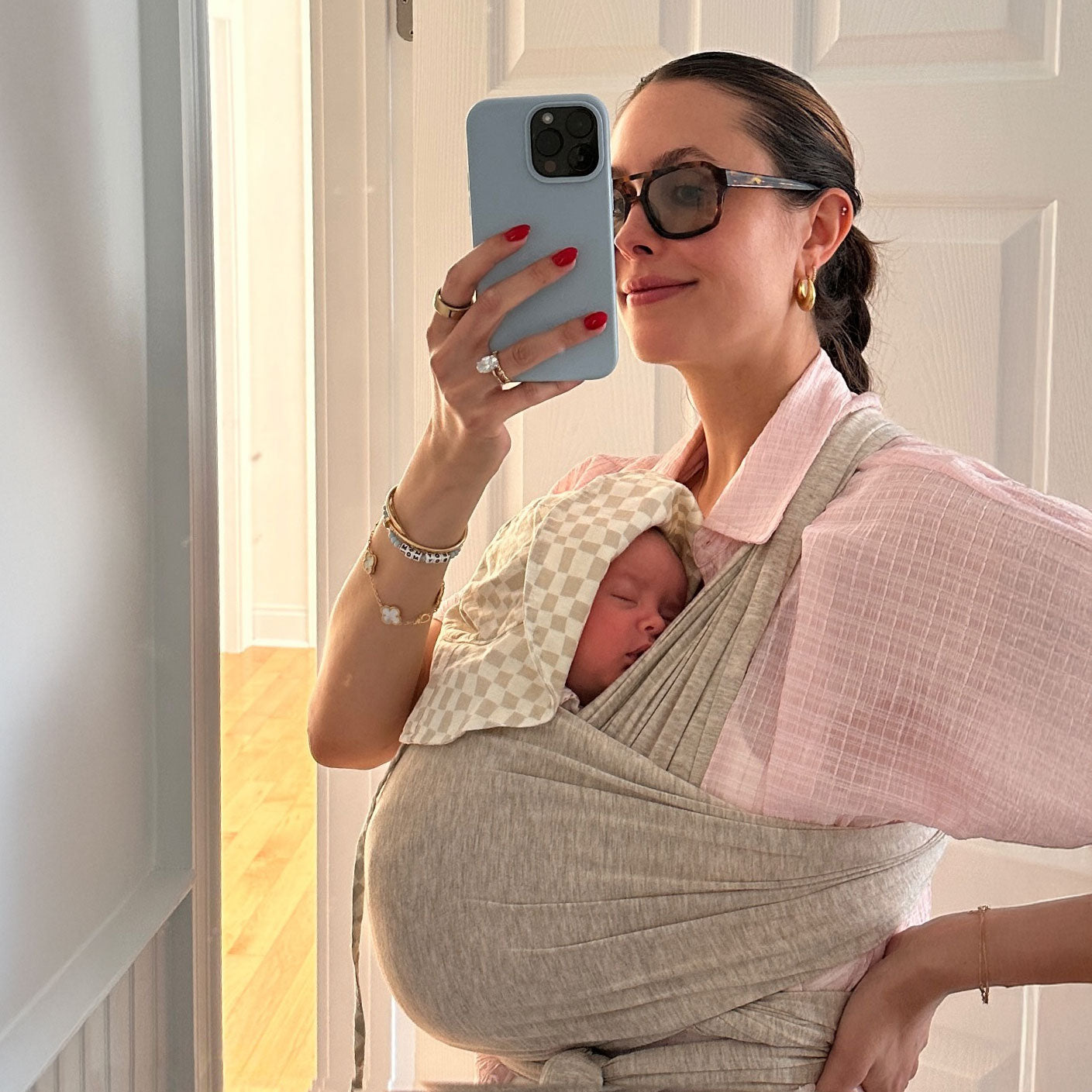 Woman in pink shirt smiles as she takes mirror selfie in bathroom while wearing baby in the Flax Solly Baby wrap