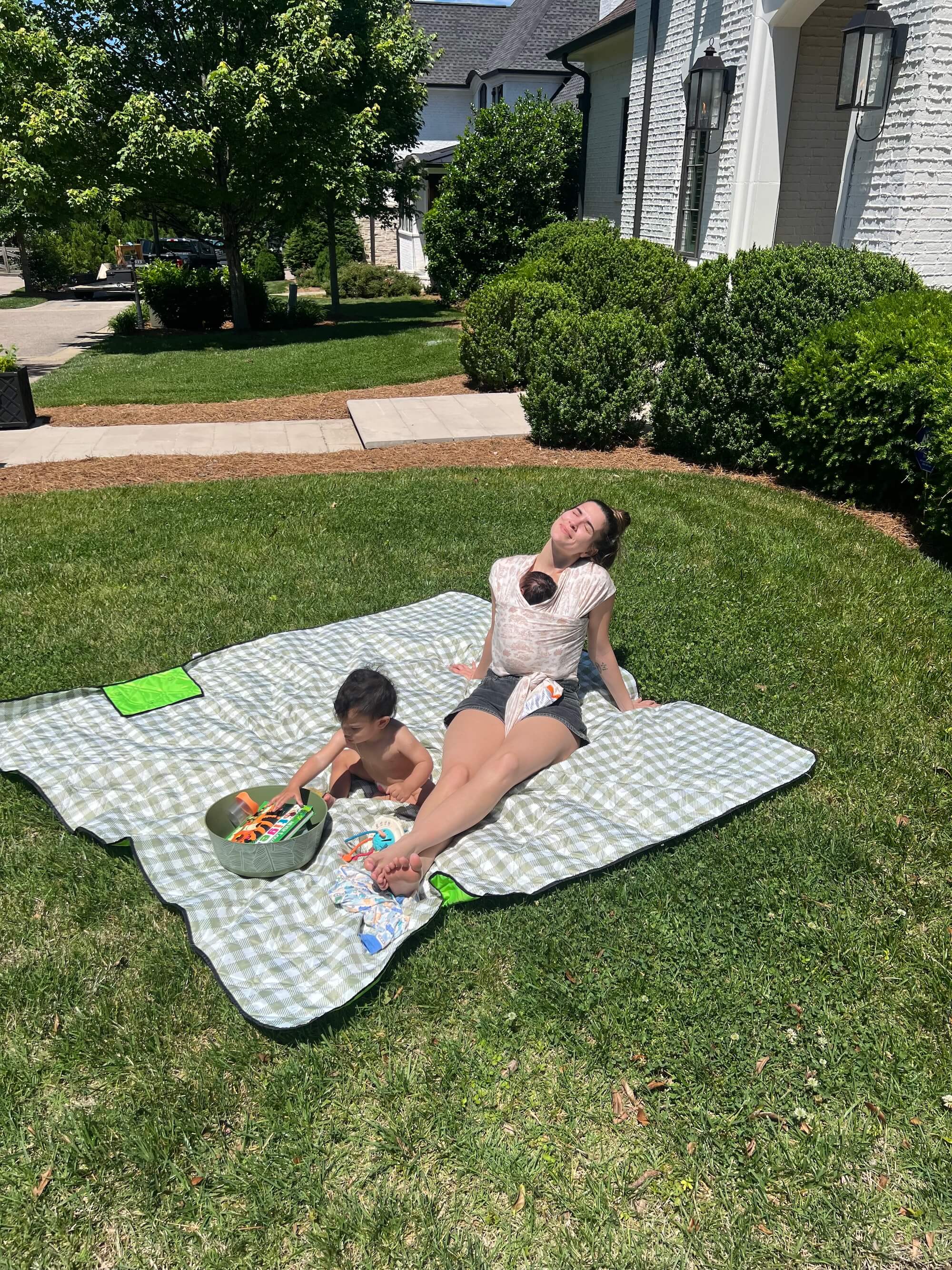 Mom sitting on blanket in yard with toddler playing and baby on her chest in a wrap carrier