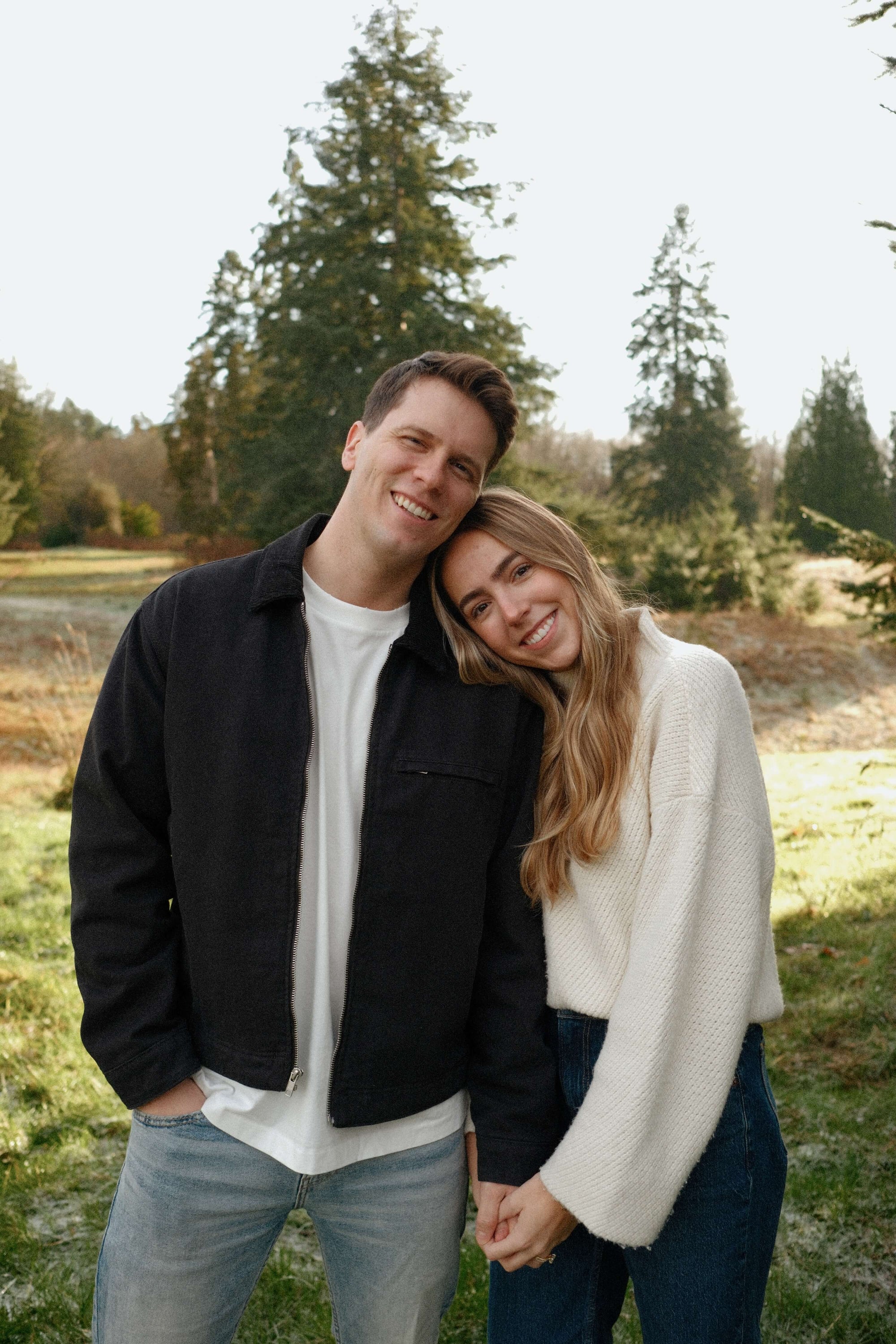 Blonde woman in cream  sweater and jeans holding hands and smiling next to her husband with trees in the background