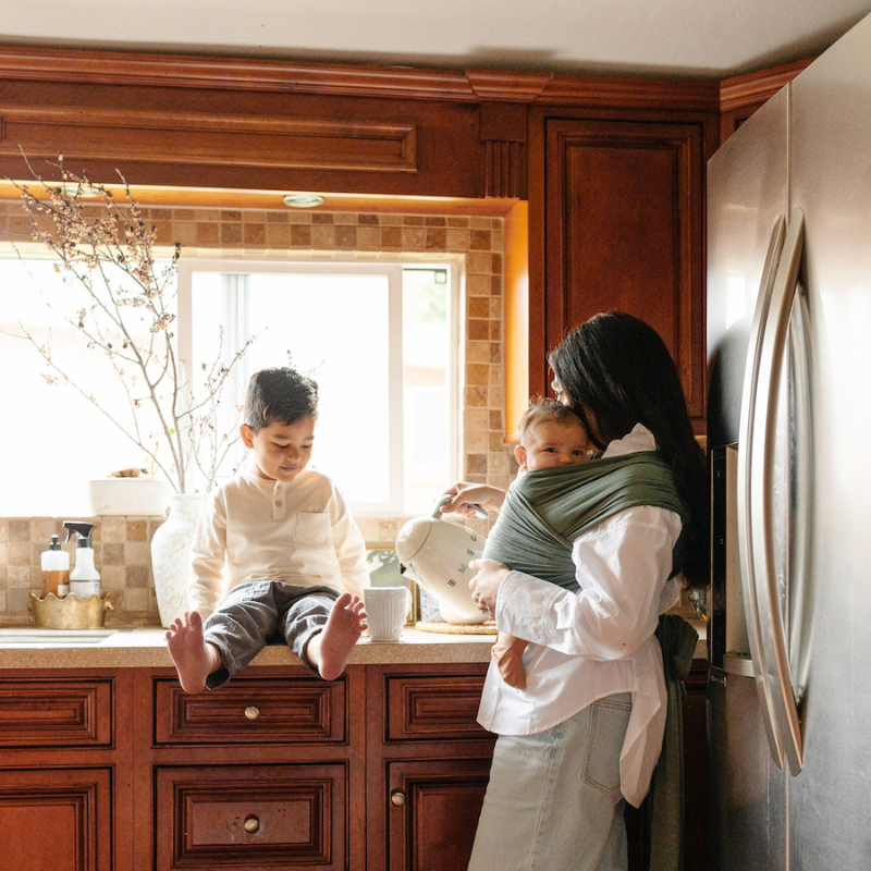 Woman pours kettle in kitchen wearing baby in the Basil Solly wrap with her toddler son sitting on the counter
