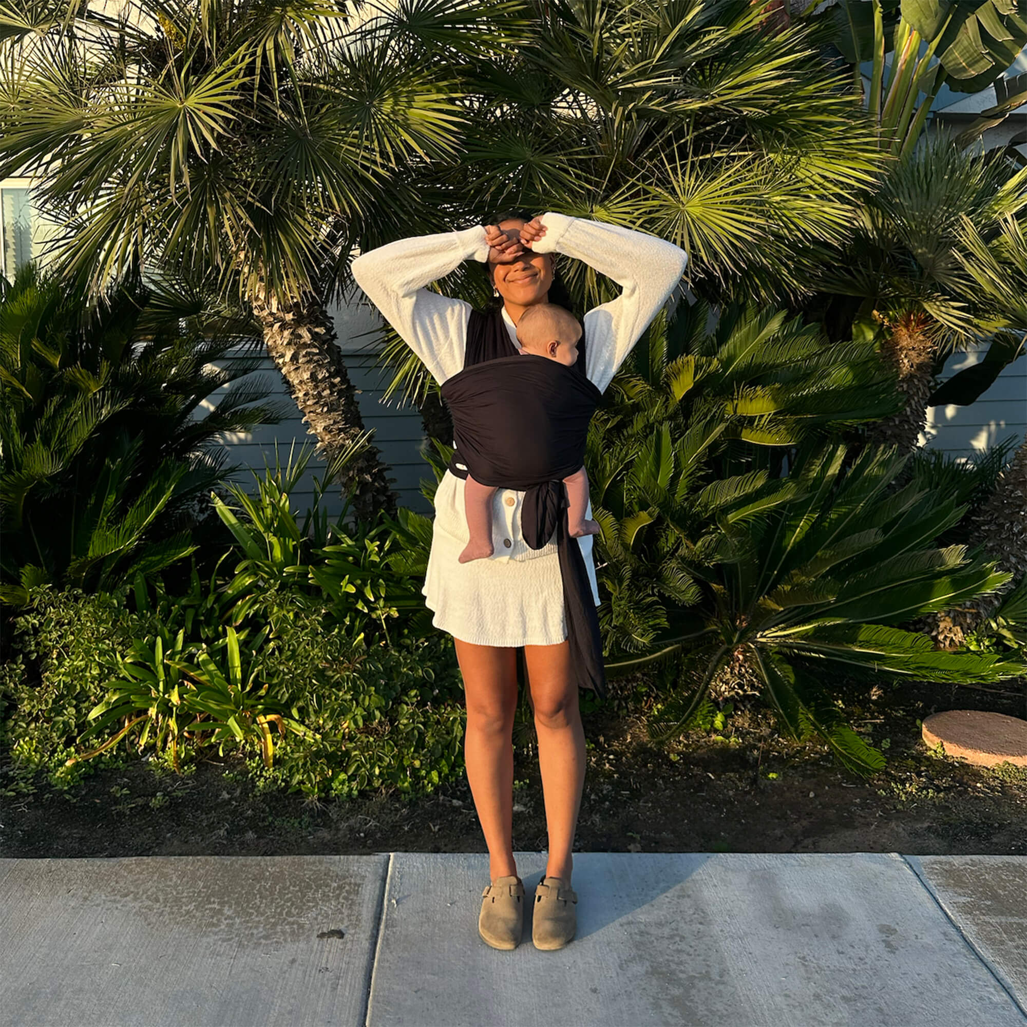 Woman stands on sidewalk in front of palm trees and smiles with hands covering her eyes from the sun. She is holding her baby in the Black Solly wrap carrier.