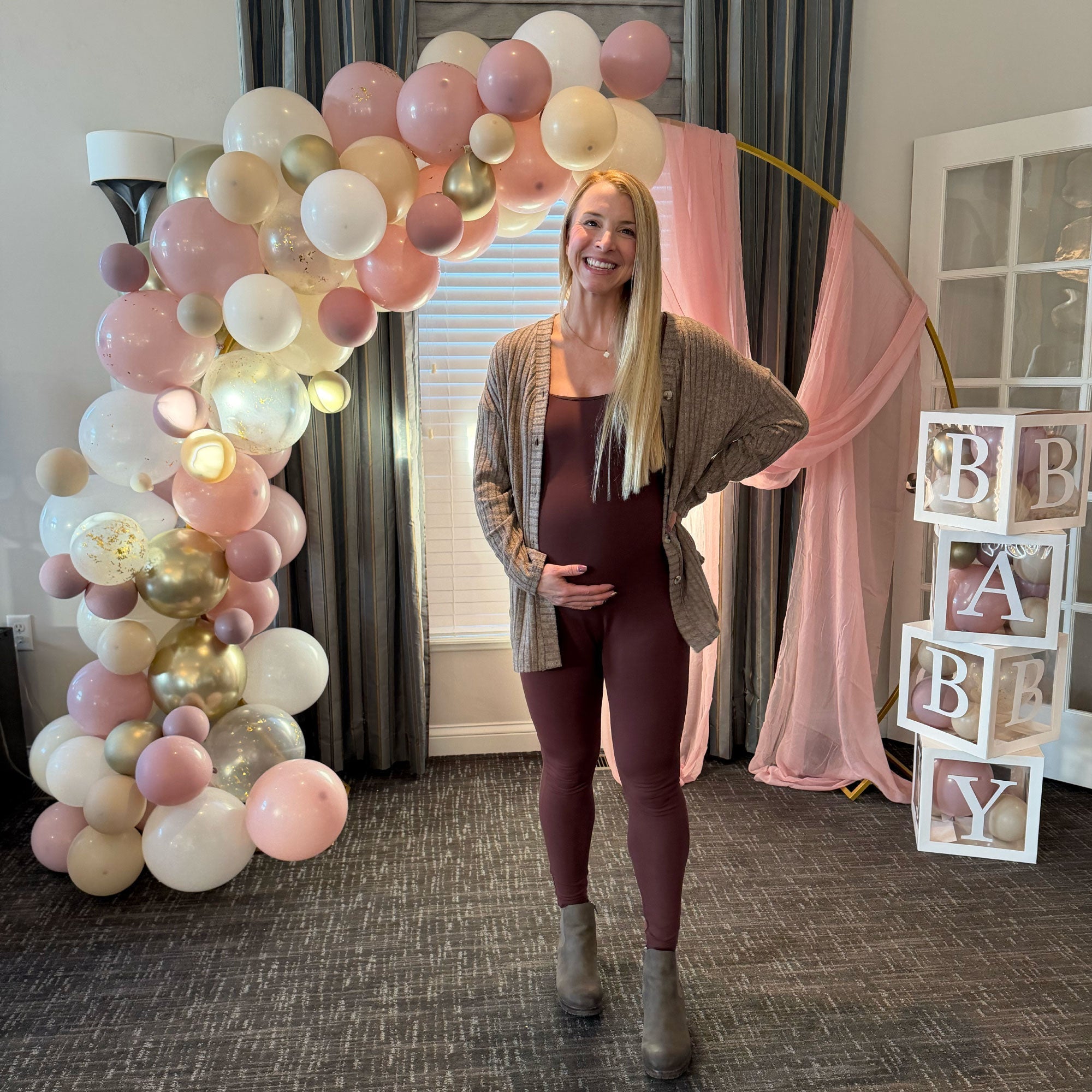 Pregnant woman smiles with hand on belly in front of a pink balloon arch at her baby shower