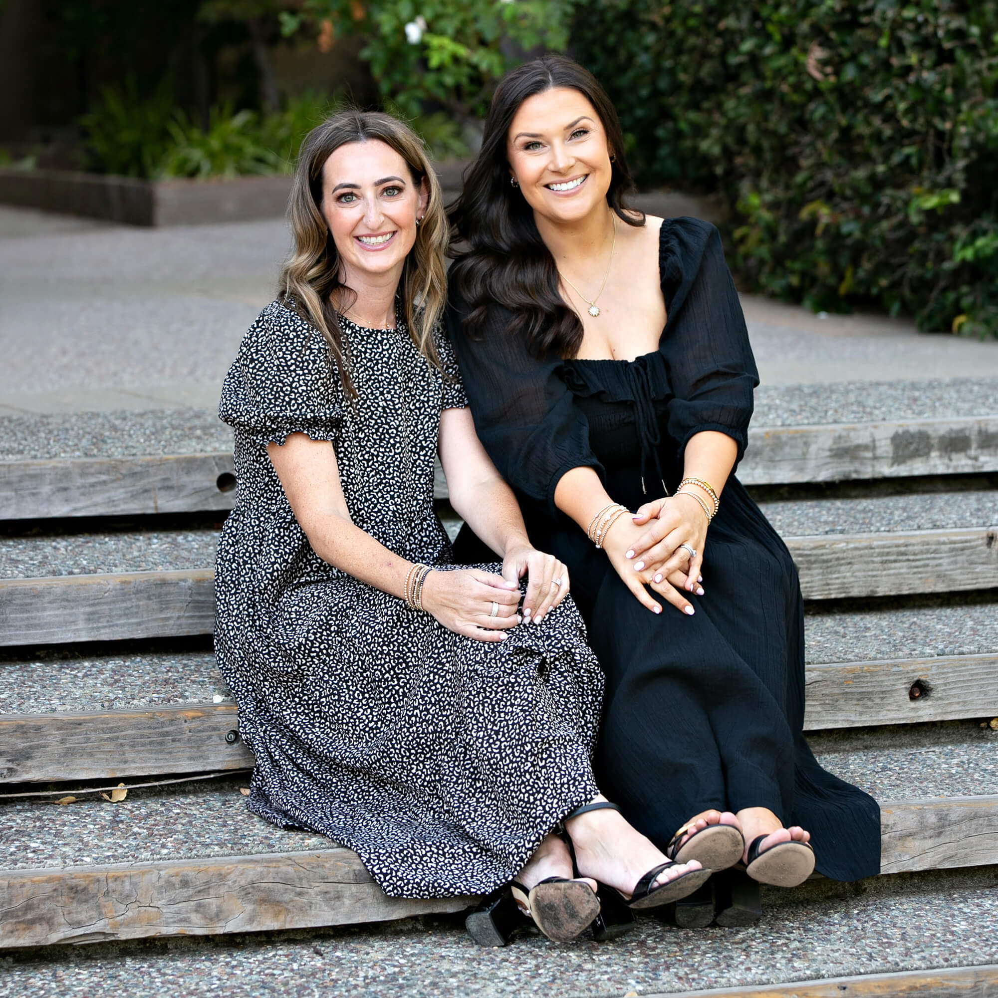 2 women in black dresses smile, seated next to one another on outdoor steps