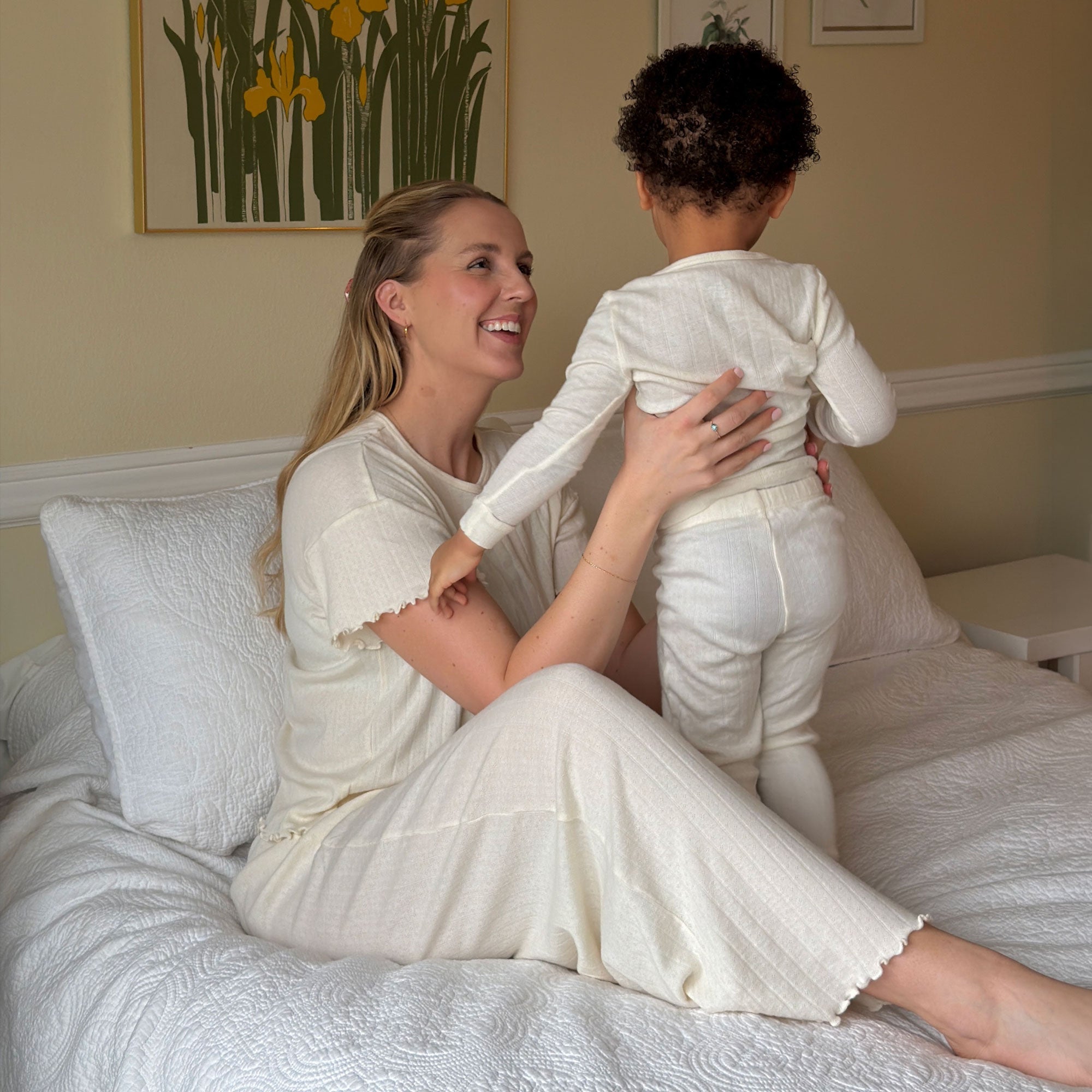 Mother sits on bed and looks lovingly up at her standing toddler in matching cream pointelle pajamas