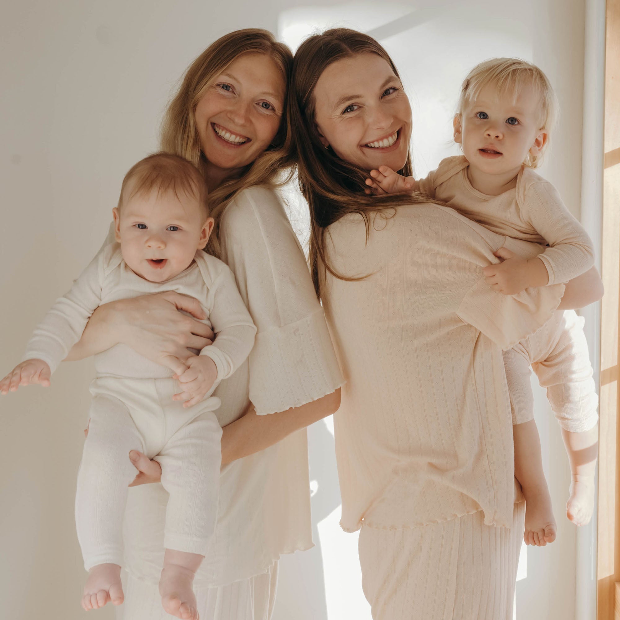 2 moms smile back to back holding their babies in matching cream and blush pointelle pajamas