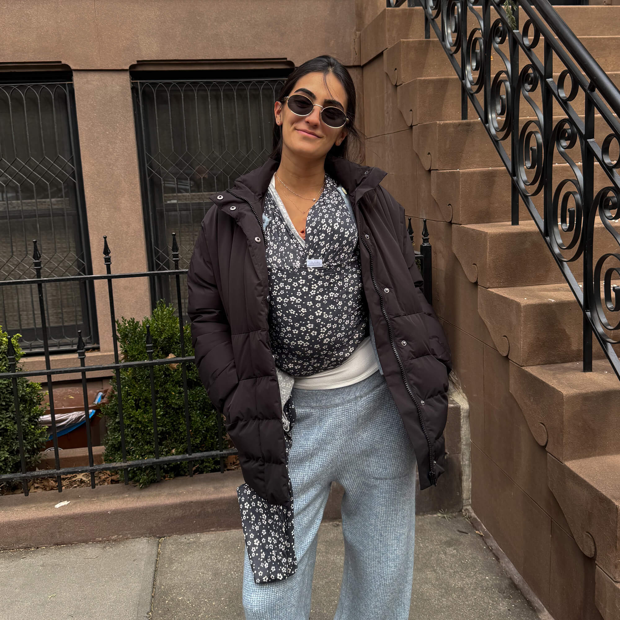 Woman stands outside NYC brownstone wearing her baby in the Solly Wrap Carrier with a brown puffy coat