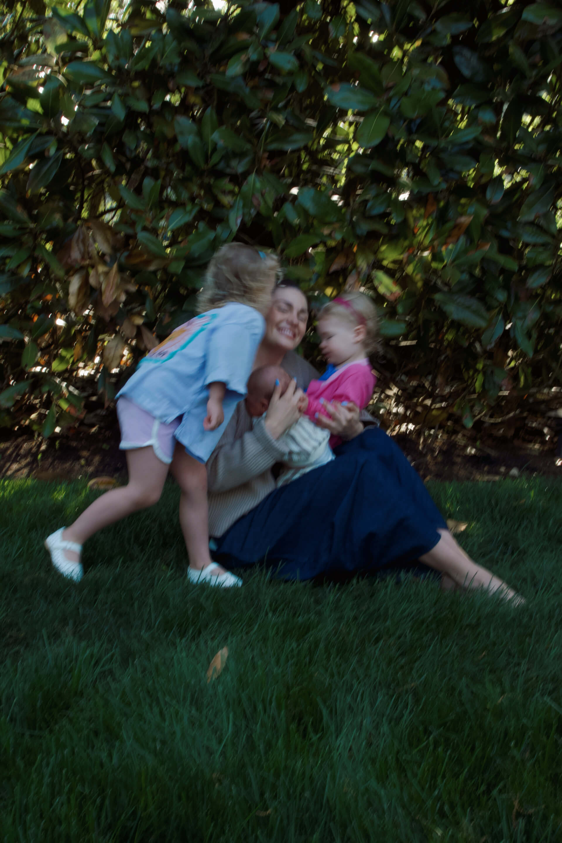 Mother sitting on grass outdoors holding a newborn while two toddlers hug her in a candid family lifestyle moment.