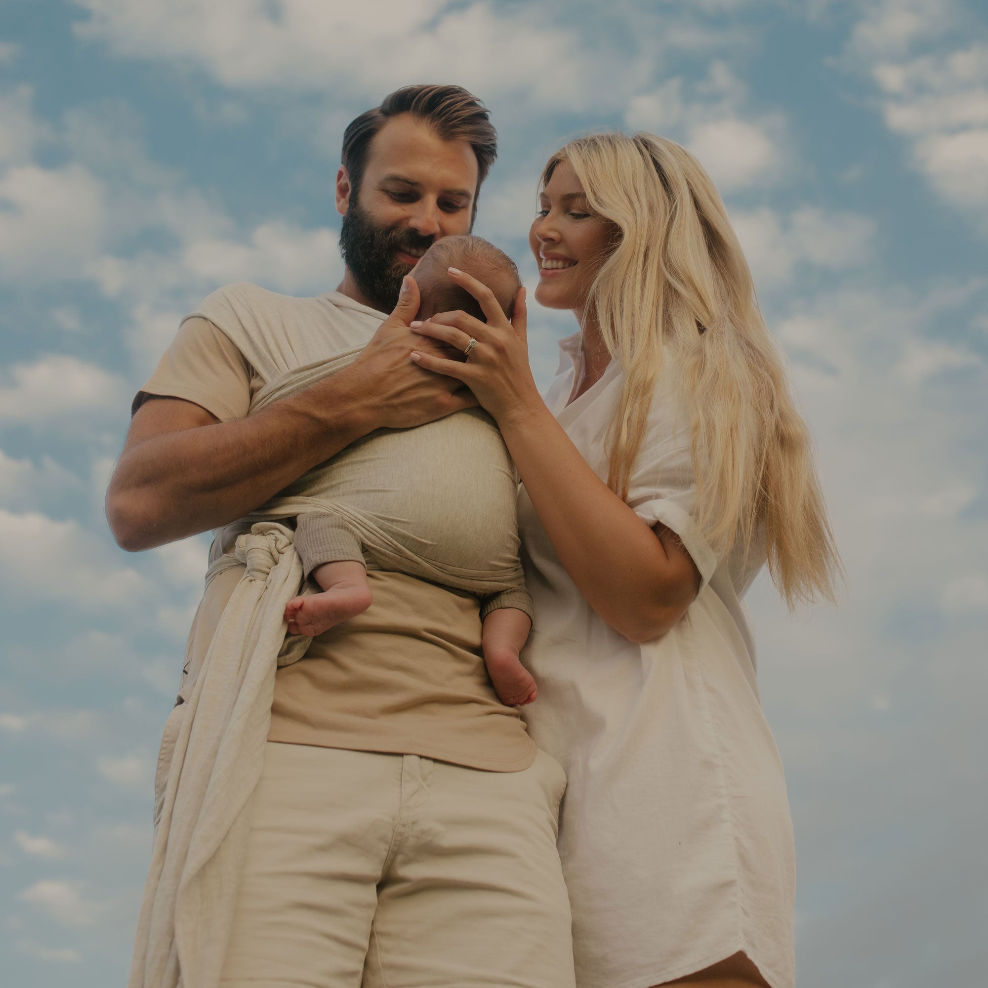 Husband holds newborn in the Flax Solly wrap and mom stands by looking lovingly at their baby with a blue sky background