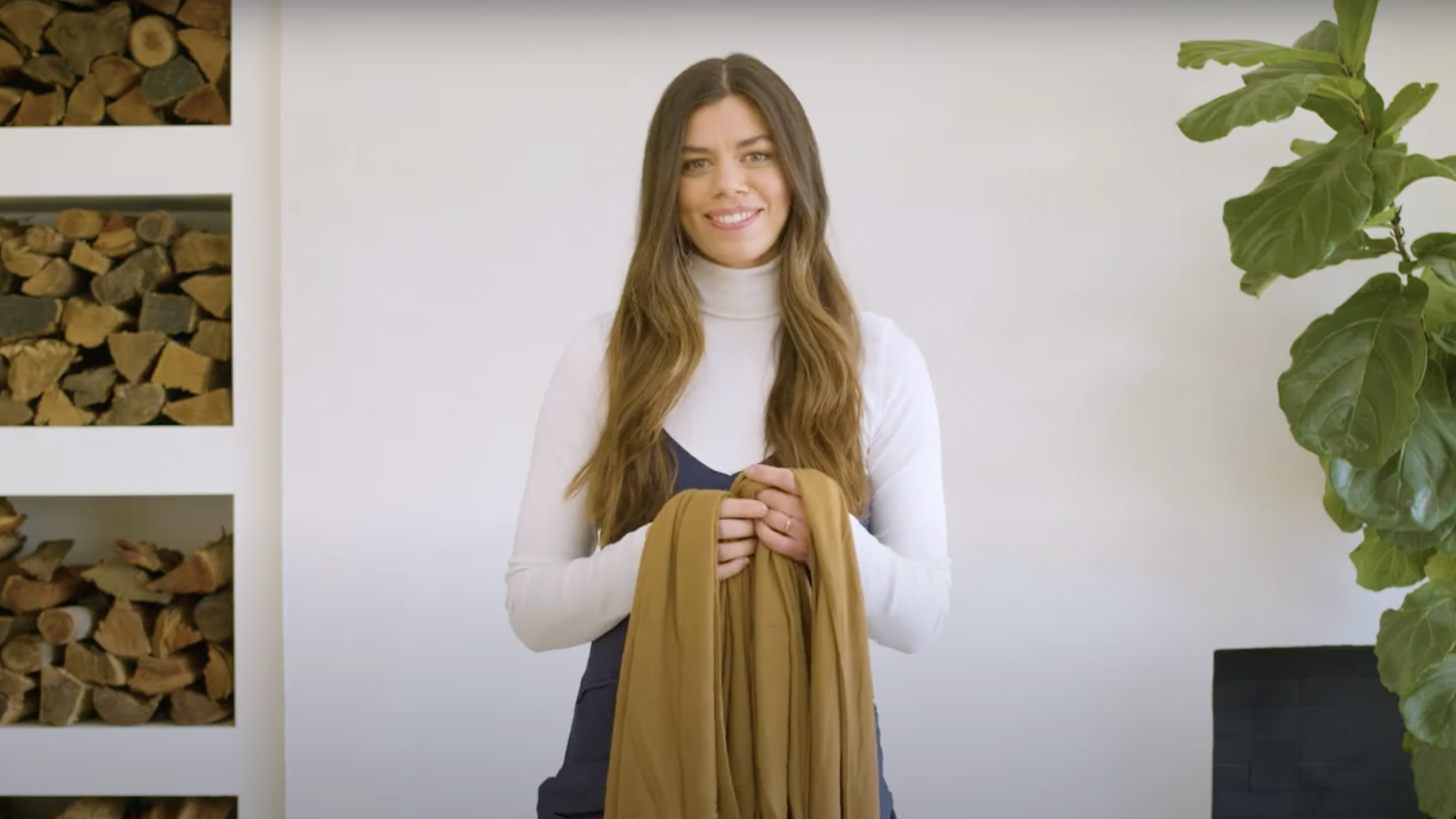 Woman wearing white turtleneck and navy dress holding mustard yellow fabric in front of white wall with wood storage and green plant.