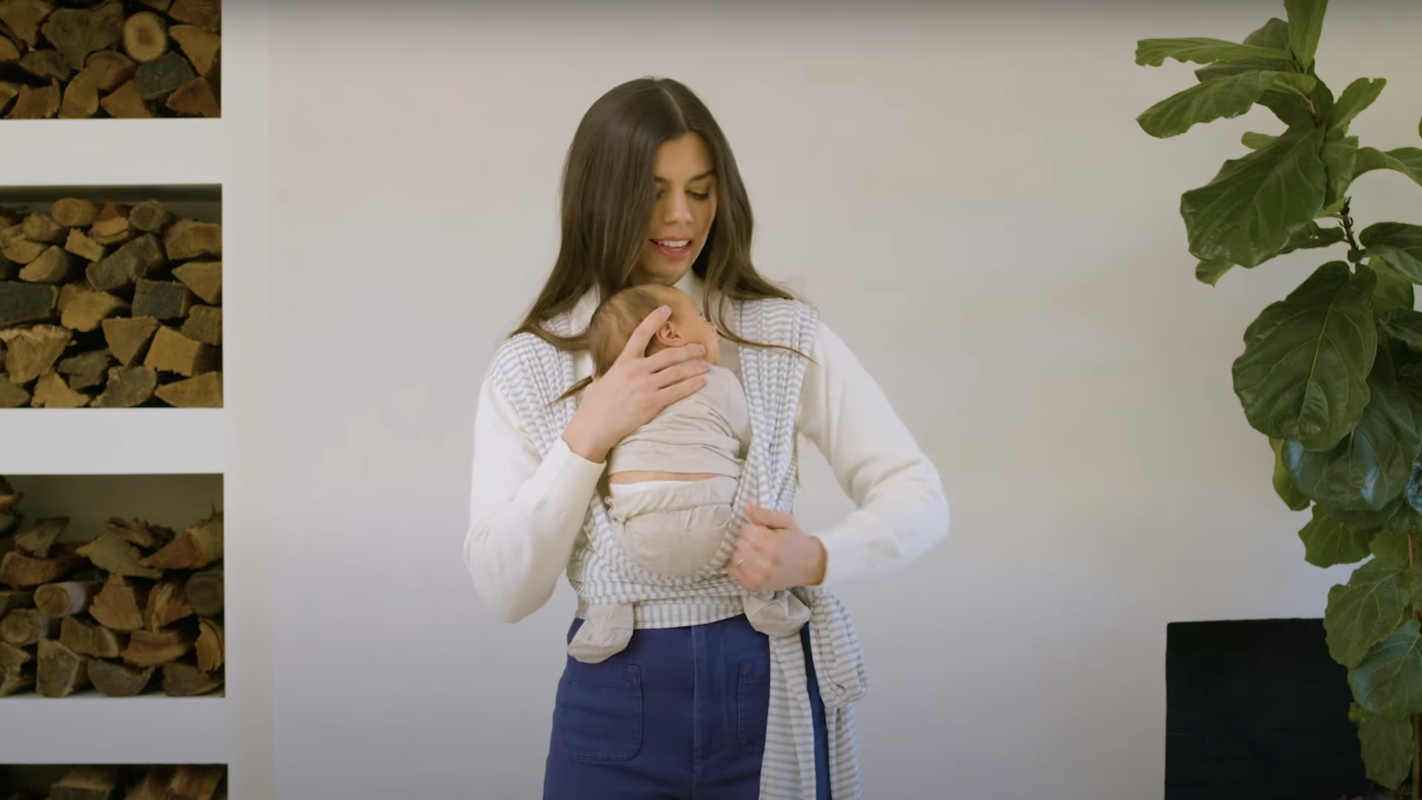 Woman wearing white sweater and blue skirt holding baby in gray and white striped baby wrap carrier indoors