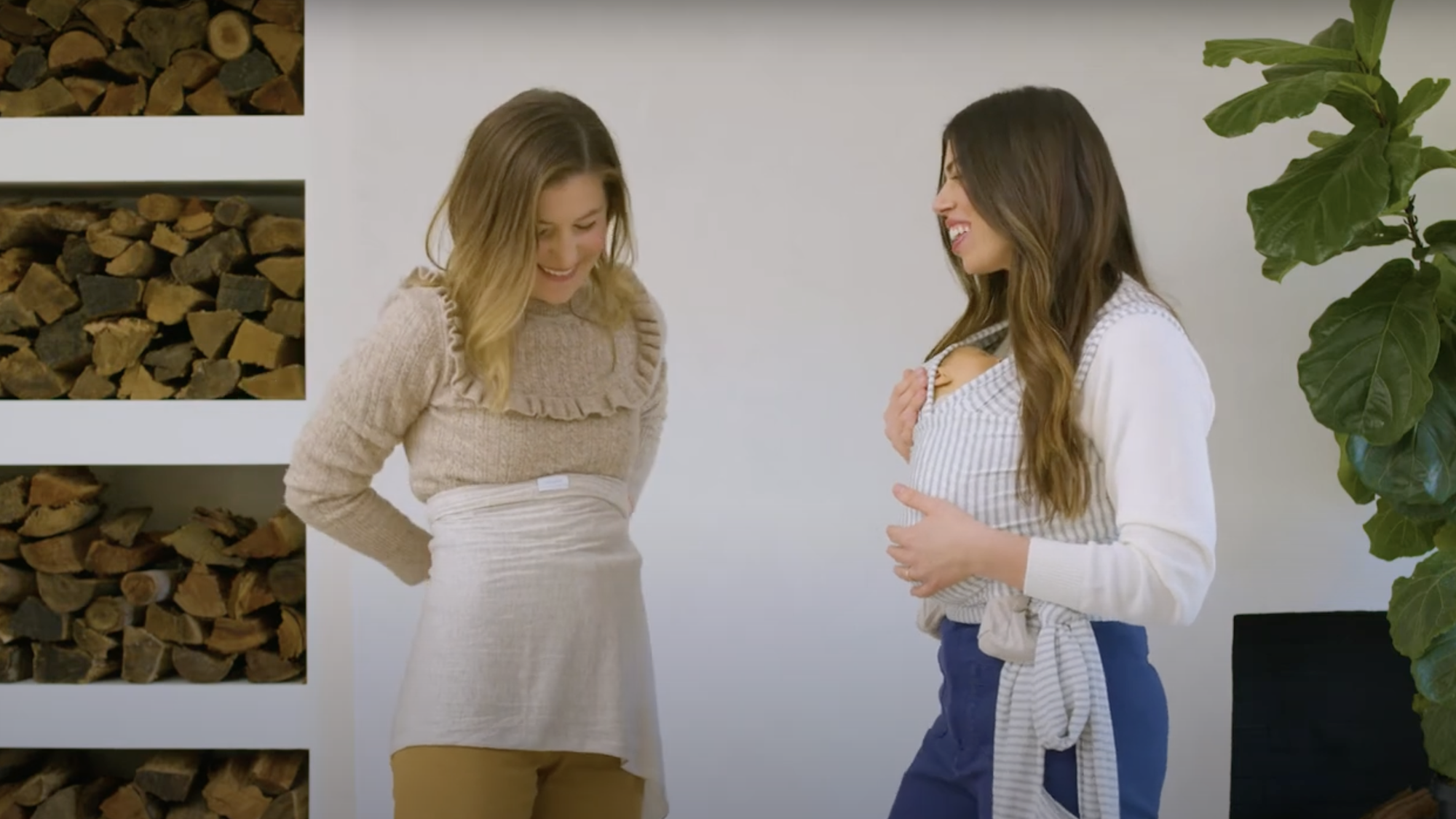 Woman wearing beige sweater and tan pants with cream-colored belly band next to woman holding baby in striped gray and white baby wrap carrier