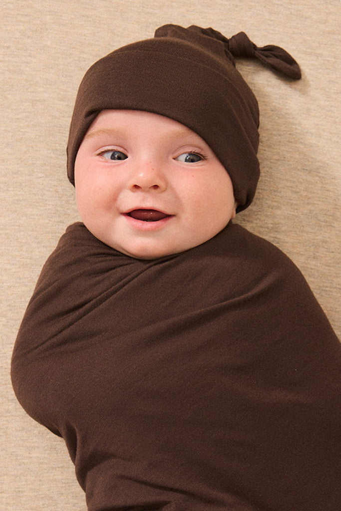 A baby wrapped in a brown swaddle blanket and wearing a matching brown knotted beanie, lying on a textured beige blanket.