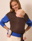 A woman wearing a brown baby wrap carrier with blue long-sleeve shirt underneath, carrying a baby against her chest, set against a plain beige background.