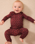 A baby wearing a dark red long-sleeve one-piece outfit with a small red floral pattern, lying on a beige textured blanket.