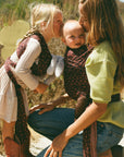 A woman and a young girl outdoors wearing matching dark red patterned baby wraps over their clothing, with the woman also carrying a baby in the wrap. The scene is set in a natural, grassy outdoor environment.