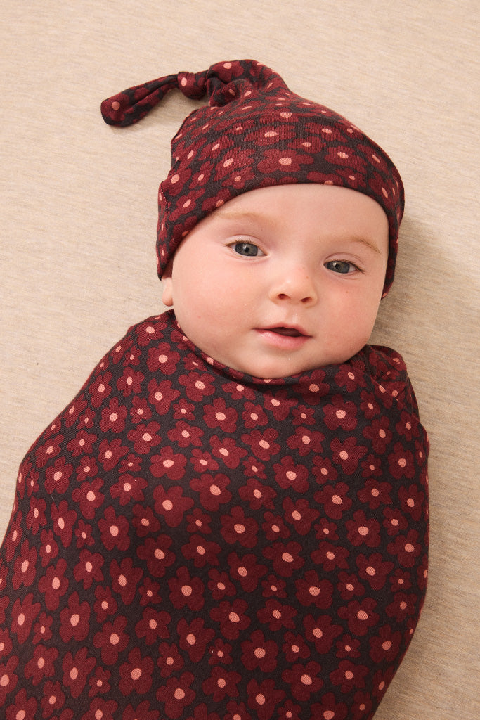 A baby swaddled and wearing a matching knit hat in a dark pink floral pattern, lying on a beige textured blanket.